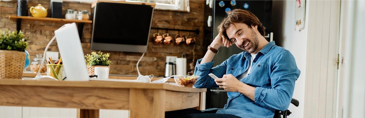 A man sits at a kitchen table with his mobile phone, smiling, relaxed, representing Ulster Bank's positive digital support for all.