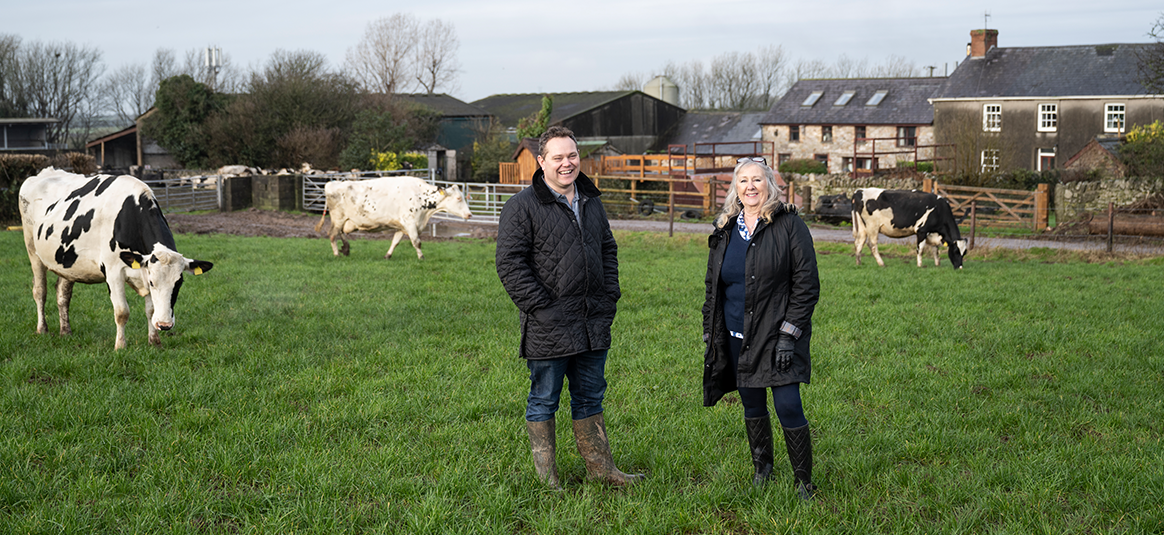 A white male and a white female farmer, smile, wearing three quarte length jackets, stand on green grass of a field where black and white dairy cows are grazing, in front of a farm house and out-buildings in the background.