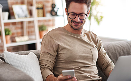 Person sitting on a couch using a laptop and holding a smartphone.
