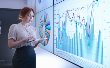 Woman holding a tablet standing in front of several wall mounted monitors showing charts