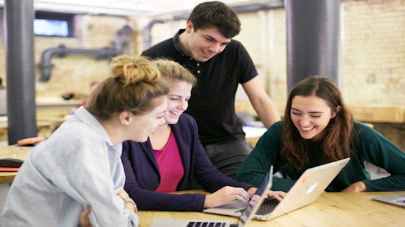 Four office colleagues, two men and two women, collaborating while gathered around a laptop in a modern office setting